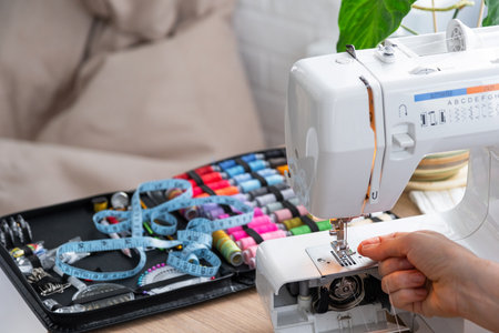 Woman hand close up sews tulle on electric sewing machine. Filling the thread into the sewing needle, adjusting the tension. Comfort in the house, a housewife's hobby, layout of sewing toolsの写真素材