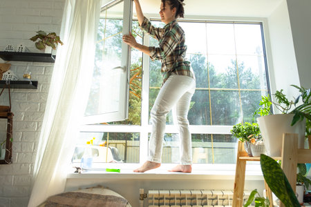 Woman manually washes the window of the house with a rag with spray cleaner and mop inside the interior with white curtains. Restoring order and cleanliness in the spring, cleaning serviceの写真素材
