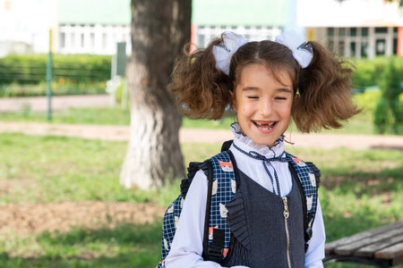 Cheerful funny girl with a toothless smile in a school uniform with white bows in school yard. Back to school, September 1. Happy pupil with a backpack. Primary education, elementary class.の写真素材