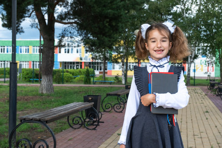 Girl with backpack, school uniform with white bows and stack of books near school. Back to school, happy pupil, heavy textbooks. Education, primary school classes, September 1の写真素材