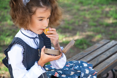Girl with backpack eating sandwich packed in a sandwich box near school. A quick snack with a bun,...