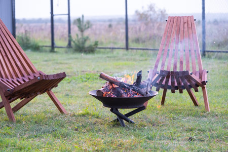 Metal fire bowl with burning wood in the yard and garden rack deck chairs. Safe decorative fire pit, Picnic area in the gardenの写真素材