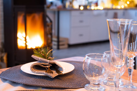 Serving festive table with plates, forks, knives, napkins, glasses close-up near stove fireplace in interior of loft house decorated for Christmas and New Year. Waiting for guests for festive dinnerの写真素材
