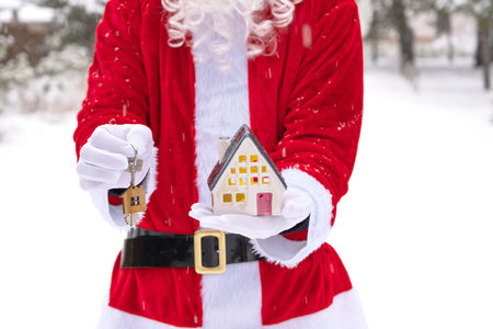 Santa carefully holds in his hands tiny house in small size and key with a keychain in snow outdoor - a gift for Christmas, New year, dream. Protection, insurance, mortgage, purchase of housingの写真素材