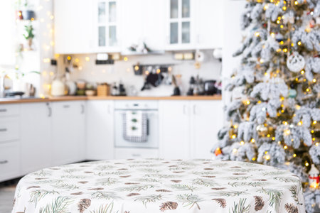 Round empty table to demonstrate the product with tablecloth with pine needles and cones print, space for text in white kitchen, modern interior with a Christmas tree. New Year, Christmasの写真素材