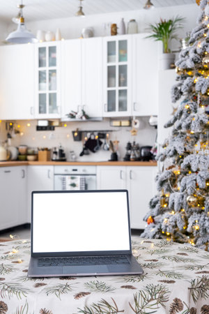 Laptop with a white screen mock up in the cozy white decorated Christmas kitchen with fairy lights and a Christmas tree. Seasonal remote work, internet, shopping, buying gifts Christmas and New Year.の写真素材