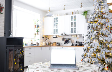 Laptop with a white screen mock up in the cozy white decorated Christmas kitchen with fairy lights and a Christmas tree. Seasonal remote work, internet, shopping, buying gifts Christmas and New Year.の写真素材