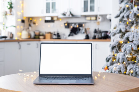Laptop with a white screen mock up in the cozy white decorated Christmas kitchen with fairy lights and a Christmas tree. Seasonal remote work, internet, shopping, buying gifts Christmas and New Year.の写真素材