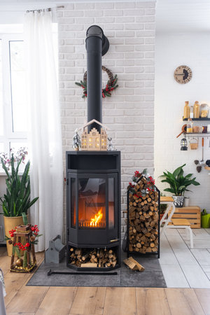 Metall black stove, burning hearth fireplace in white Festive interior of house is decorated for Christmas and New Year, Christmas tree. firewood in the woodpile, cozy and heating of homeの写真素材