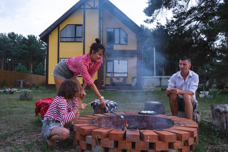 Family mom, dad, daughter roast marshmallows on skewers on a campfire in the hearth in the courtyard...