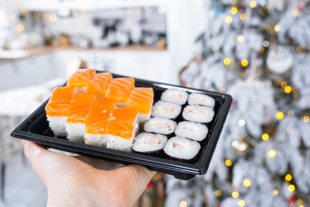 Delivery of sushi and rolls set is held in hand in the festive interior of house with a white Christmas tree for Christmas and New year. Food delivery for the holidaysの写真素材