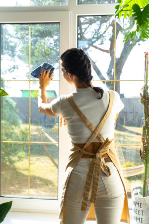 Woman in apron manually washes the window of house with rag cleaner and mop inside the interior with...