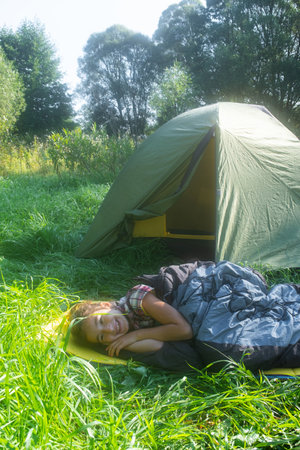 The girl is lying in a sleeping bag on a warm mat near a tent outdoor. Family camping recreation, hiking equipment.の写真素材