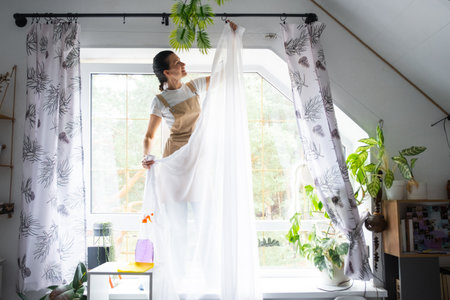 Woman in apron hangs transparent tulle curtains on large attic windows in the house inside the interior with potted plants. Spring cleaning, tidying upの写真素材