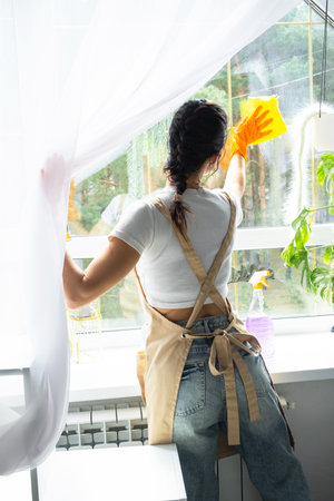 Woman in rubber gloves and apron manually washes window of house with rag cleaner and mop inside interior with home plants on windowsill. Restoring order and cleanliness in spring, cleaning serviceの写真素材