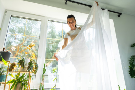 Woman in apron hangs transparent tulle curtains on large windows in the house inside the interior with potted plants. Spring cleaning, tidying upの写真素材