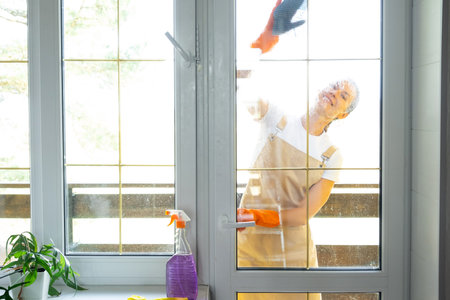 Woman in rubber gloves and apron manually washes window balcony of house with rag cleaner and mop...