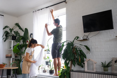 Husband helps his wife hangs transparent tulle curtains on large windows in the house inside the interior with potted plants. Spring cleaning, tidying upの写真素材