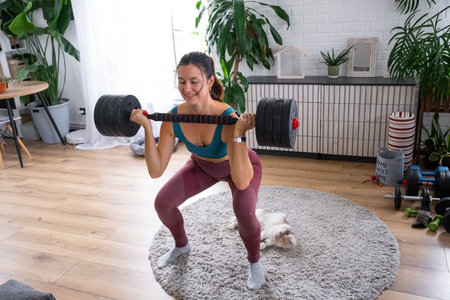 A woman in a sports uniform is engaged in fitness and strength training with a barbell and dumbbells at home in the interiorの写真素材