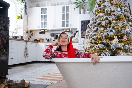 Happy woman in Santa hat is lying resting in New bathtub under Christmas tree as New Year's gift in interior of decorated kitchen. Interior renovation and design, construction and home improvementの写真素材