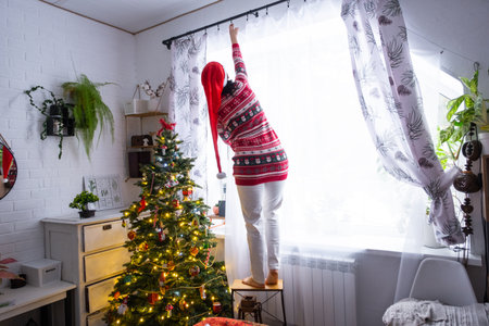 A woman in a Santa hat hangs curtains in the bedroom on the skylights with a Christmas tree and Christmas decorations, preparing for the New Yearの写真素材