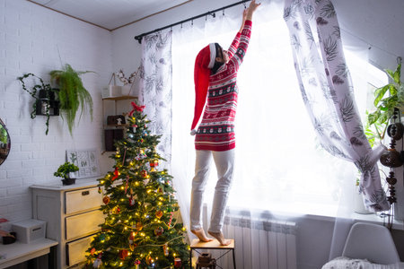 A woman in a Santa hat hangs curtains in the bedroom on the skylights with a Christmas tree and Christmas decorations, preparing for the New Yearの写真素材