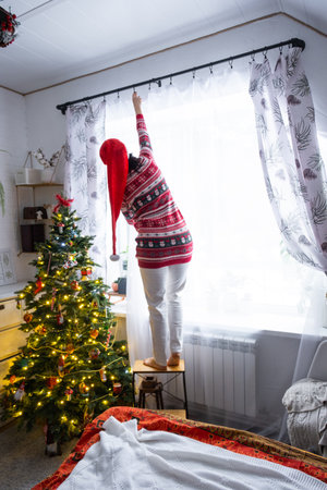 A woman in a Santa hat hangs curtains in the bedroom on the skylights with a Christmas tree and Christmas decorations, preparing for the New Yearの写真素材
