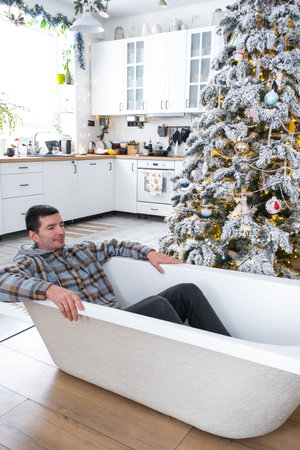Man is lying resting in bathtub under Christmas tree in interior of white decorated kitchen....