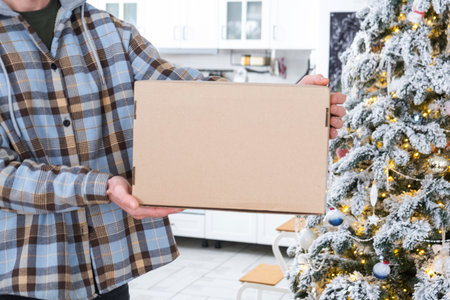 A courier with a parcel box in the kitchen with white Christmas decorations and a Christmas tree. Gift delivery for New Year and Christmas, courier serviceの写真素材