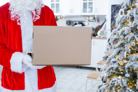 Courier Dressed as Santa with parcel box in the kitchen with white Christmas decorations and a Christmas tree. Gift delivery for New Year and Christmas, courier serviceの写真素材