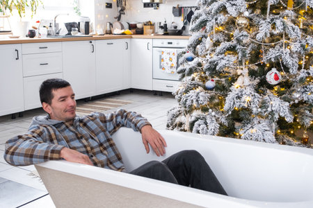 Man is lying resting in bathtub under Christmas tree in interior of white decorated kitchen. Interior renovation and design, construction and home improvementの写真素材