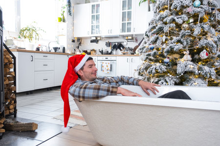 Man in Santa hat is lying resting in New bathtub under Christmas tree as New Year's gift in interior of white decorated kitchen. Interior renovation and design, construction and home improvementの写真素材