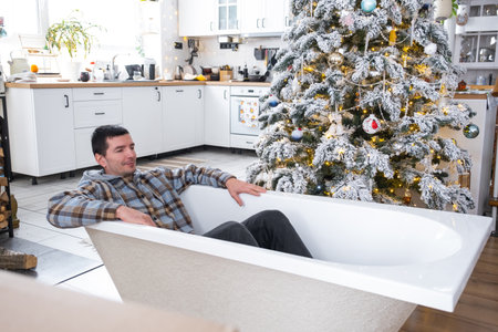 Man is lying resting in New bathtub under Christmas tree as New Year's gift in interior of white decorated kitchen. Interior renovation and design, construction and home improvementの写真素材