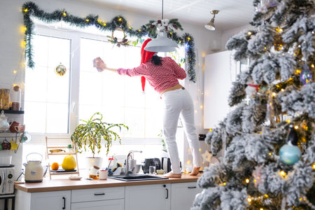 A woman in a Santa hat cleans a kitchen window with Christmas decorations, preparing for the New Year, decorating and tidying up the houseの写真素材