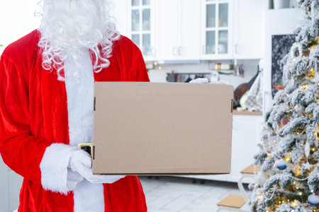 Courier Dressed as Santa with parcel box in the kitchen with white Christmas decorations and a Christmas tree. Gift delivery for New Year and Christmas, courier serviceの写真素材