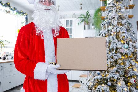 Courier Dressed as Santa with parcel box in the kitchen with white Christmas decorations and a Christmas tree. Gift delivery for New Year and Christmas, courier serviceの写真素材