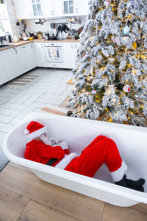 Santa is lying resting in bathtub under festive Christmas tree as gift in interior of white modern decorated kitchen. Interior renovation and design, construction and home improvementの写真素材