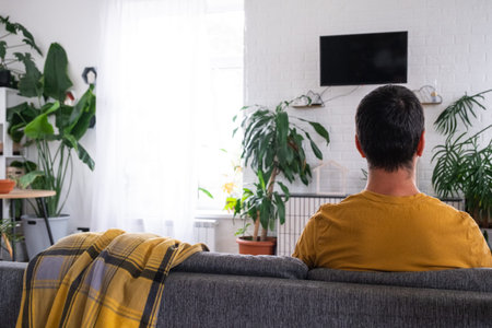 man at home in a white interior sits on the couch and watches TV, black screen mock up. Home...