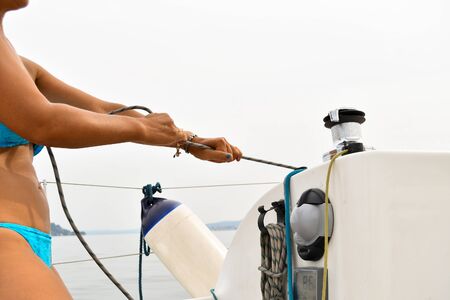 Closeup of the hands with blue nail polish of a female skipper in a blue swimsuit pulling a rope while the sailboat goesの写真素材