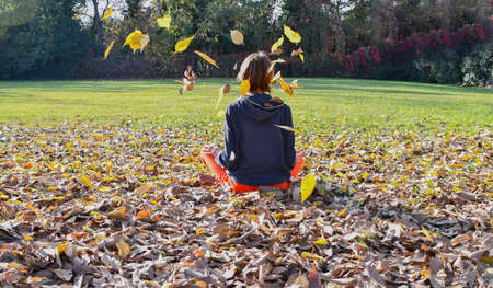 Female in sportswear sitting on a bed of autumn leaves and some falling on her head at the park on a sunny dayの写真素材