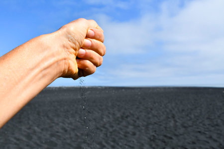 Closeup of a closed hand dropping grains of black volcanic sand with clearing and cloudy sky as a...