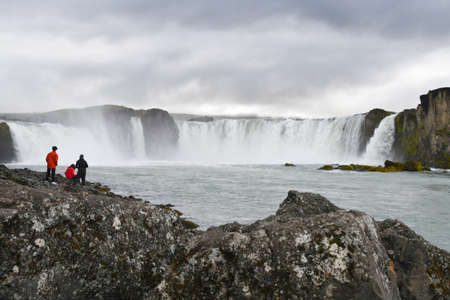 Amazing waterfall with plenty of water and three tourists who admire it enchanted during a cloudy dayの写真素材