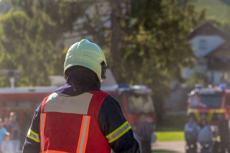 A firefighter in a suit and helmet photo in the back as he goes into action. Smoke in the background of people and fire trucks.の写真素材