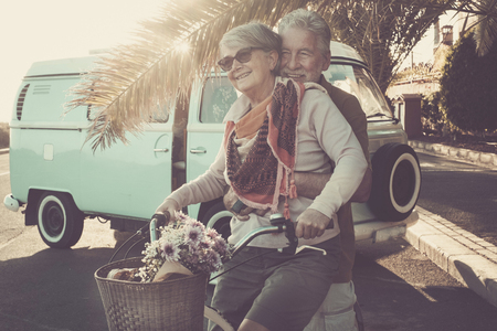 two senior aged couple go bike together in traveler vacation activities. playing and live a funny lifestyle. sun in backlight and old vintage van.の写真素材
