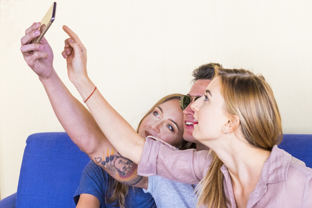 3 guys sitting on a blue sofa take pictures with phone. Two women with blonde long hair. The boy with sunglasses and tattoosの写真素材