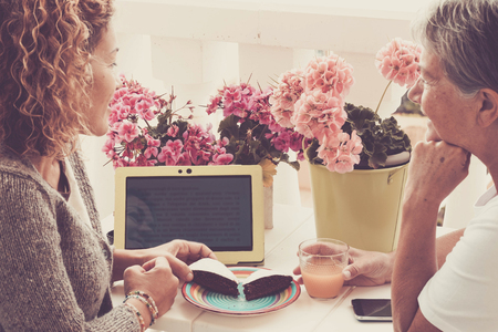 couple of friends women middle and third age 40 and 70 year old stay together in happy leisure afternoon eating some cake and drinking fruits. reading something on laptop and a mobile phone on the tableの写真素材