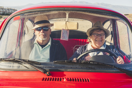 Senior man and woman with hats have fun in the summer inside an old red car. She siles and he is serious with sunglasses. Positive young old concept.の写真素材
