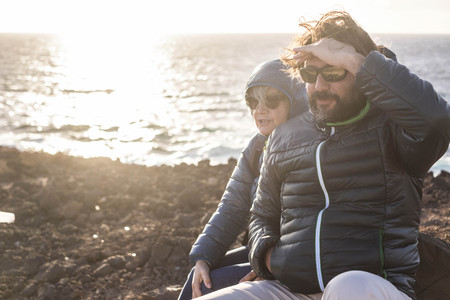 Mother and son spend time together sitting on a rock smiling and looking at sea and mountainsの写真素材