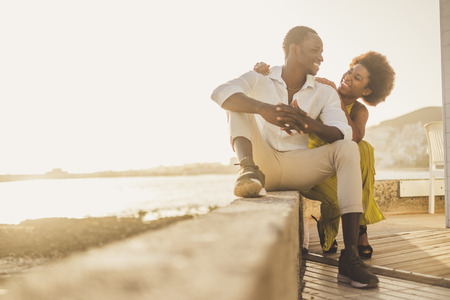 beautiful black race african couple in love and vacation sit down enjoying together with big smiles and laugh. casual clothes like fashion style with nice sunset backlight on the background. tenerife location.の写真素材