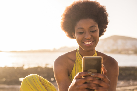 daily summer sunnny scene with black race afro american beautiful girl with alternative hair looking at the phone and check social media. sunny golden light beach and ocean in the background. vacation conceptの写真素材